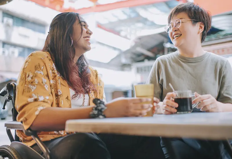 Social Support Through NDIS Two people sitting outdoors at a café table, laughing and enjoying drinks together.