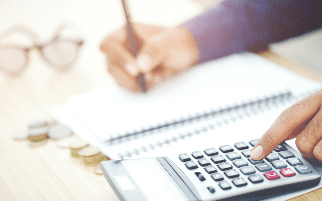 A person using a calculator while writing in a notebook, with coins and glasses on the table, representing how managing your NDIS budget categories can feel overwhelming