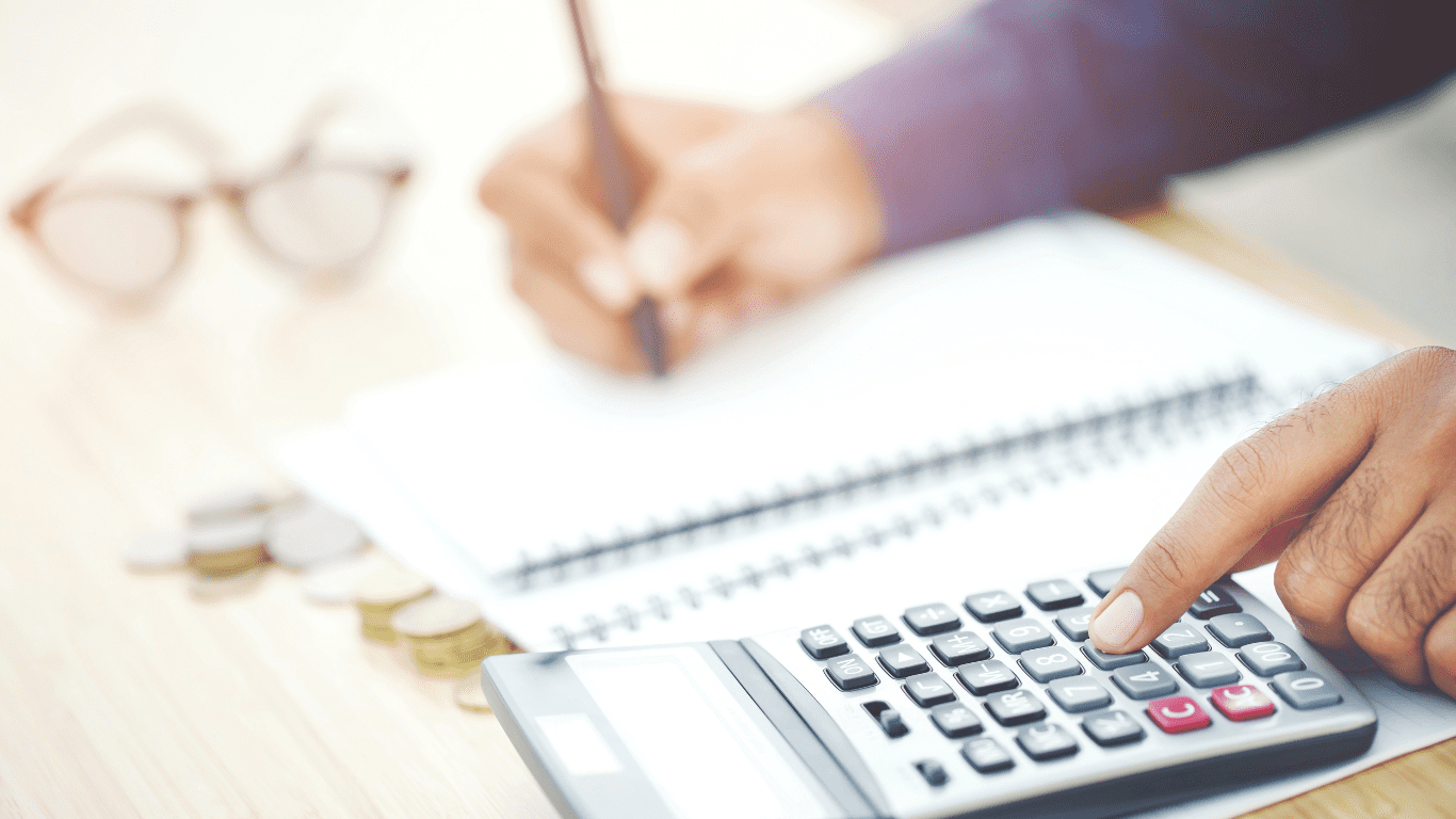 A person using a calculator while writing in a notebook, with coins and glasses on the table, representing how managing your NDIS budget categories can feel overwhelming