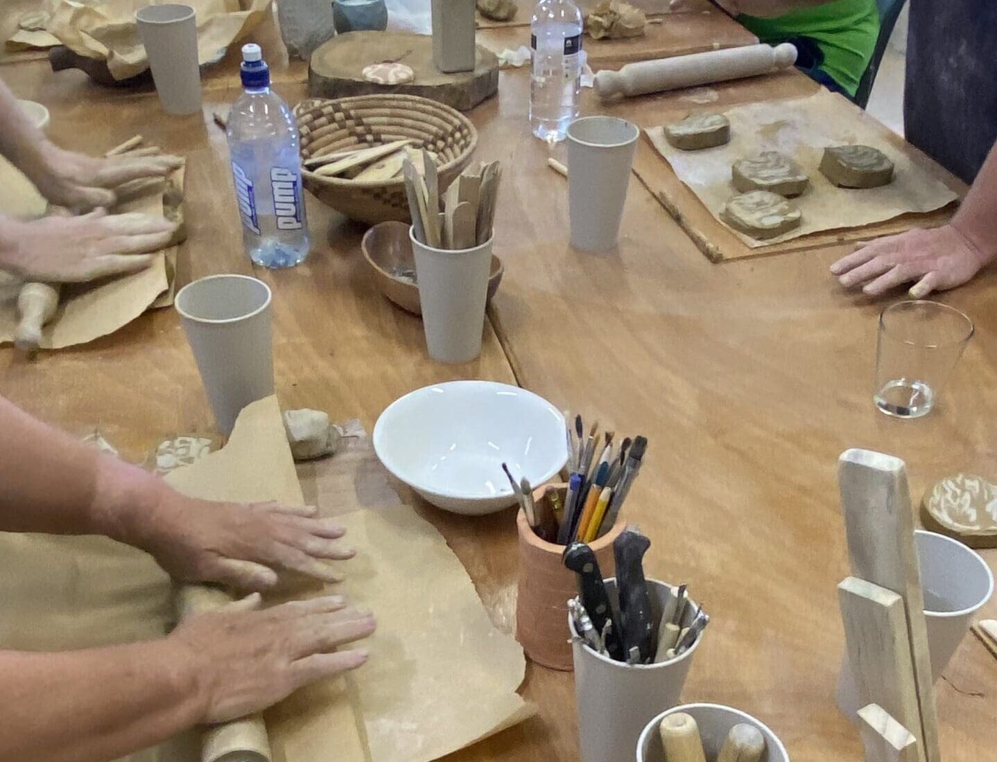 Hands shaping clay with pottery tools on a shared workshop table.