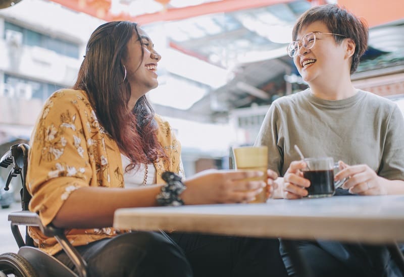 Psychosocial Recovery Coaching – Inclusive Support with Skylight Two women laughing and having coffee at an outdoor café, one using a wheelchair. Symbolising supportive and inclusive psychosocial recovery coaching.