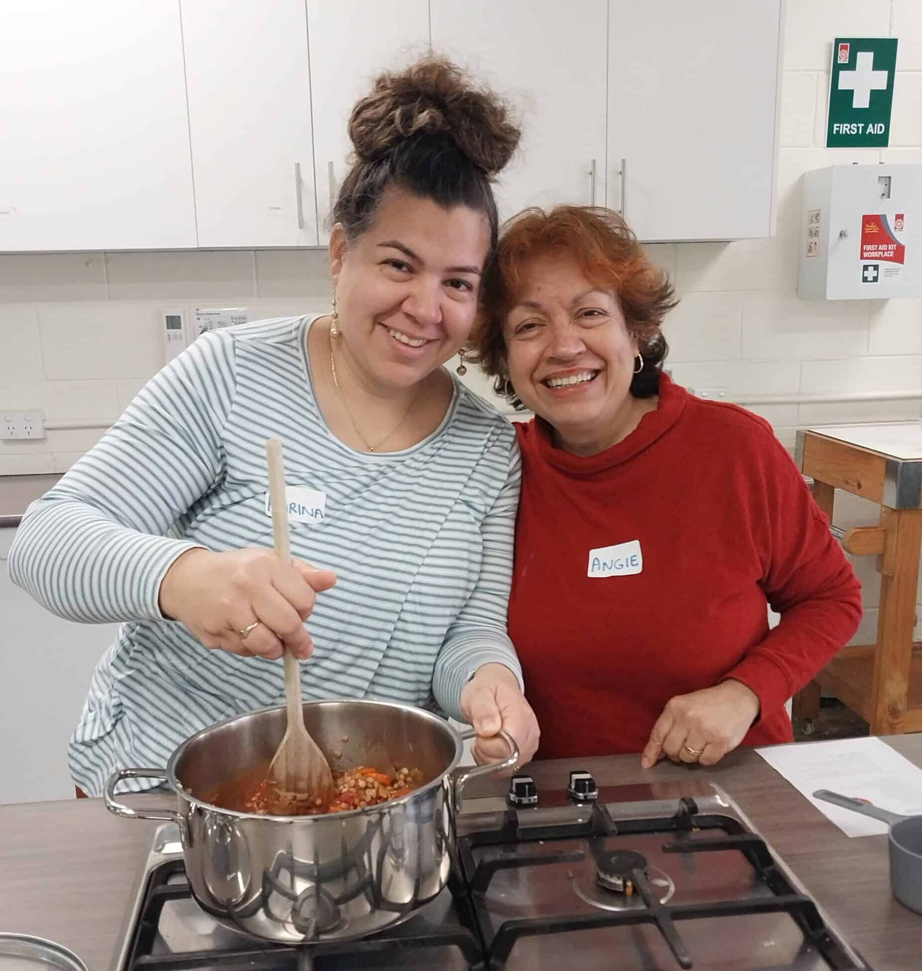 Two women smiling while cooking together at a Skylight Monday Hub session, standing at a kitchen bench with a pot on the stove.